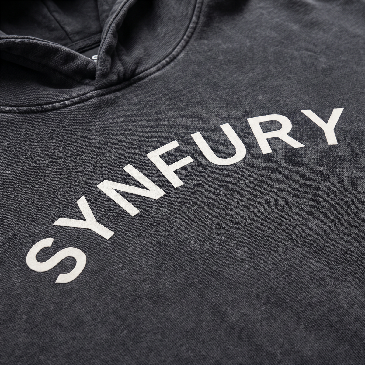 Close-up of Ash Black hoodie chest featuring SYNFURY word mark, heavyweight cotton, garment-washed finish"