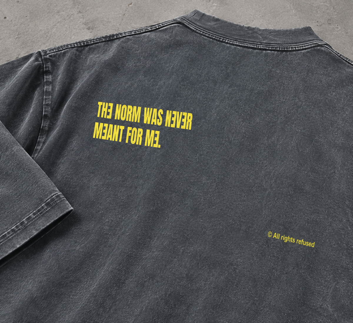 Stone Gray heavyweight tee, close-up back detail — mirrored yellow print reading 'The norm was never meant for me' with '© All rights refused' below, flat lay on concrete surface