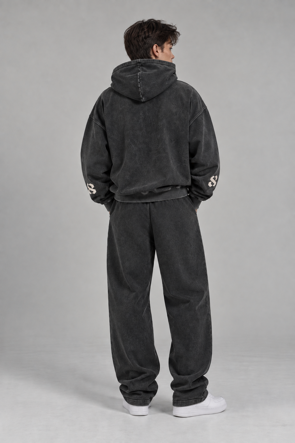 The Core Collection Set worn on model, back view — Ash Black heavyweight cotton hoodie and matching sweatpants, sleeve 'S' logo detail visible, styled with white sneakers