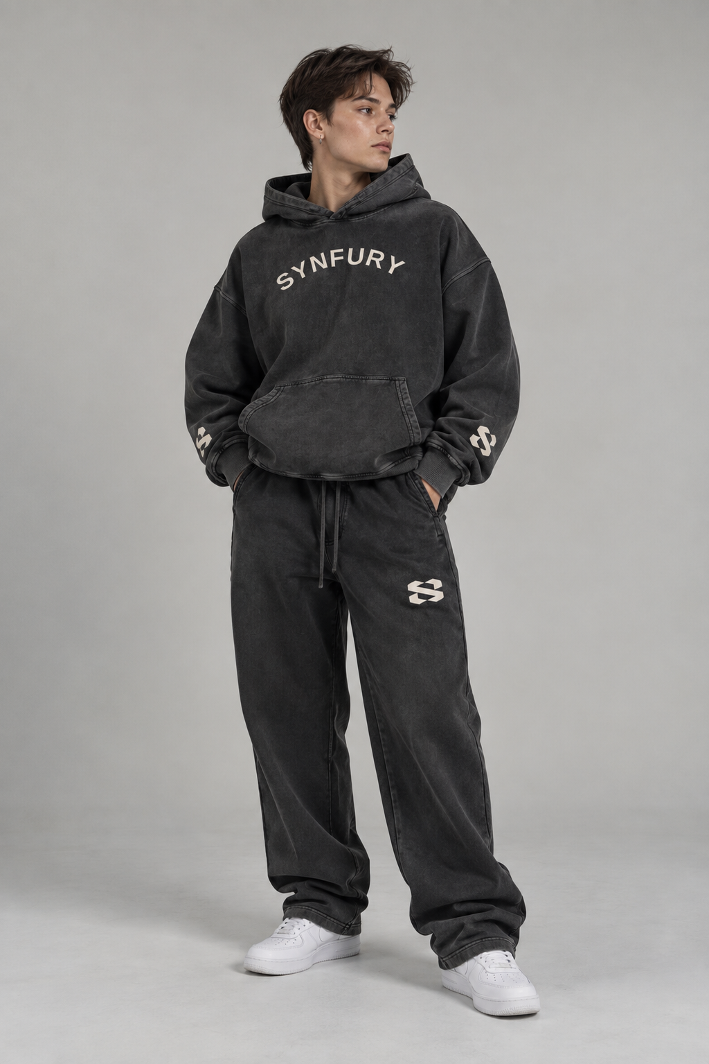 The Core Collection Set worn on model — Ash Black heavyweight cotton pullover hoodie and matching sweatpants, snow-wash finish, SYNFURY word mark and 'S' logo, styled with white sneakers