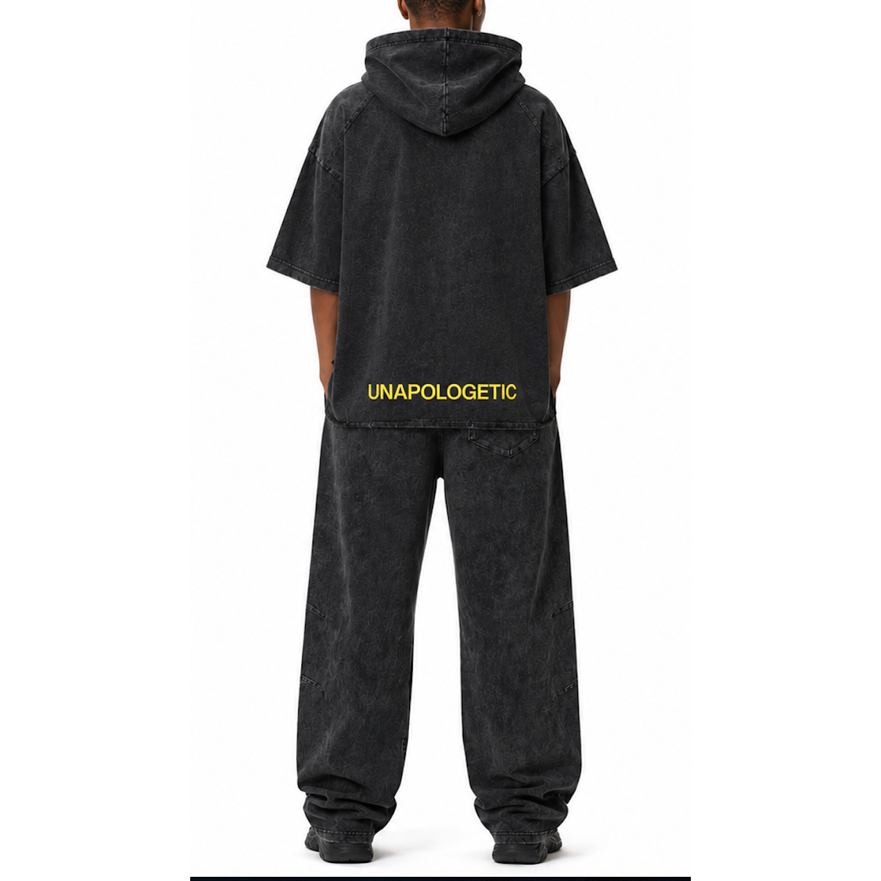 Model shown from the back wearing a matching SYNFURY Cinder Black hooded sweatshirt and wide-leg trail sweatpants set. The hoodie has 'UNAPOLOGETIC' printed in yellow across the lower back.