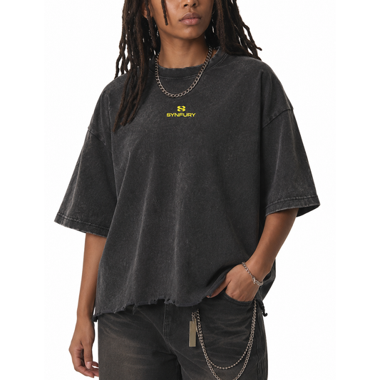A person wearing an oversized faded black t-shirt with a yellow SYNIFURY logo on the chest, paired with dark gray jeans and silver chain accessories, against a plain white background.