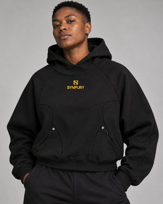 Masculine-presenting woman wearing a black SYNFURY hoodie with a yellow chest logo and curved front pockets against a light gray background.